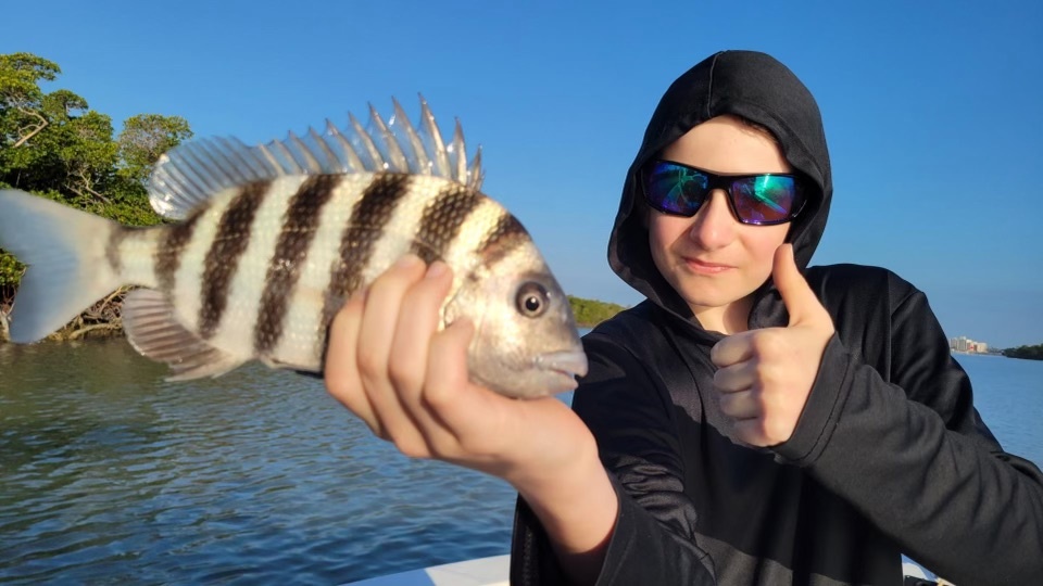 Person in hoodie holding a fish and giving a thumbs up on a boat.