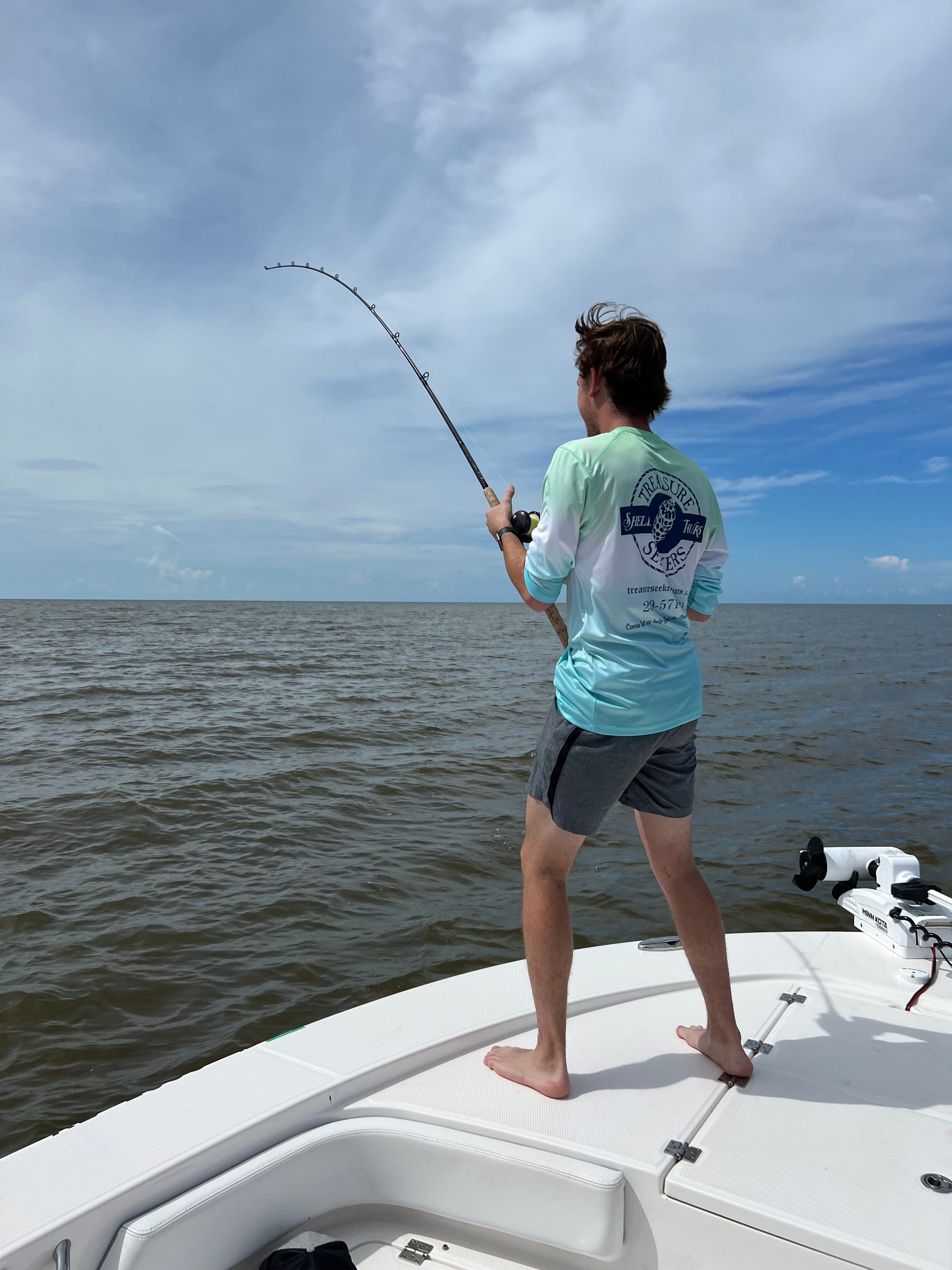 Person fishing from a boat, standing barefoot, wearing a blue shirt and shorts.