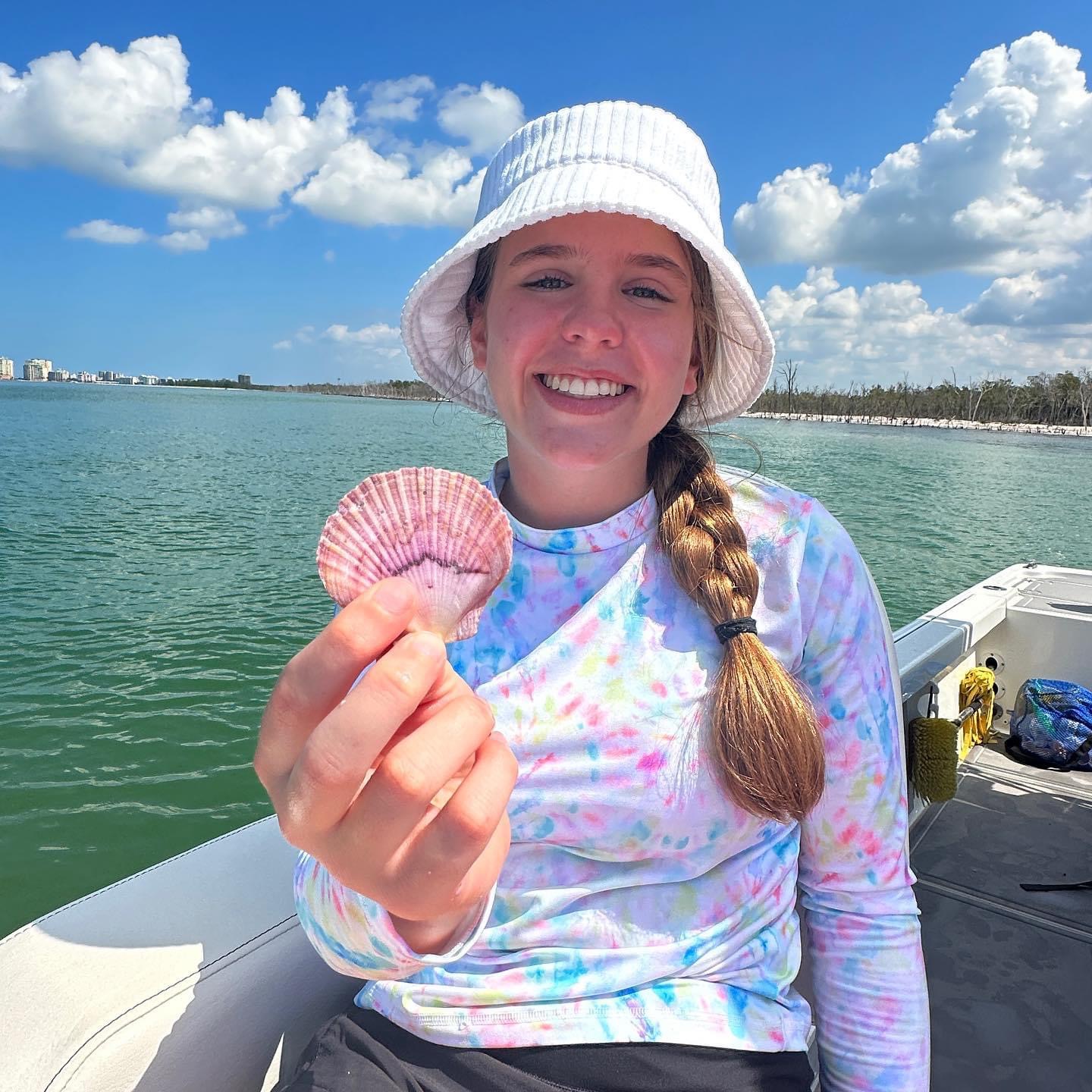 Smiling person on a boat holding a seashell, wearing a tie-dye shirt and white hat.