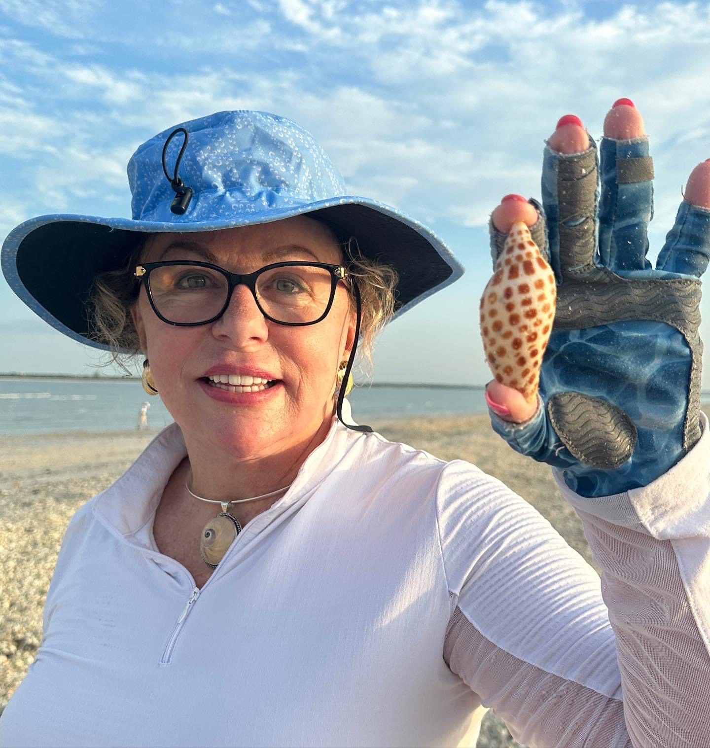 Woman in hat and gloves holding a spotted shell on a beach.