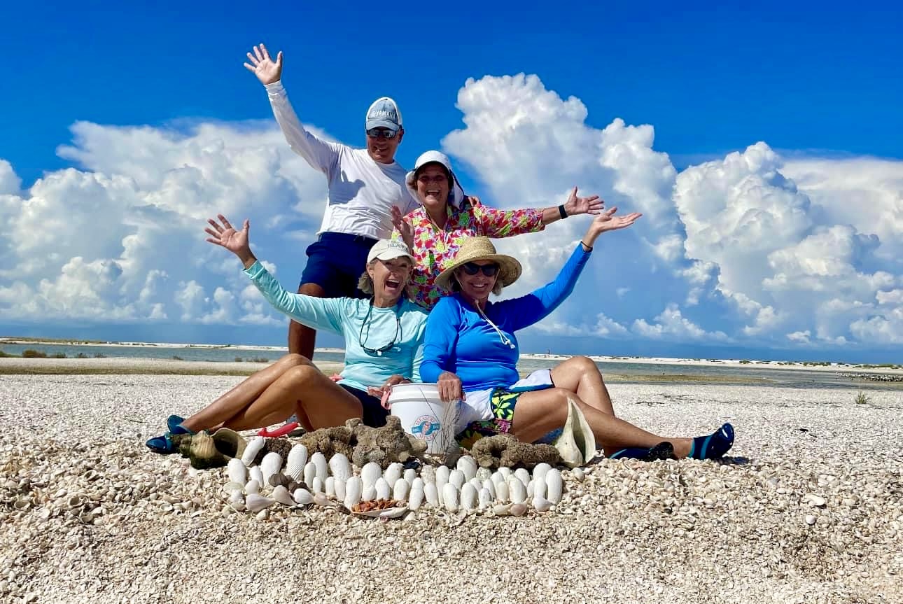 Four people posing on a beach with seashells, cloudy sky background.