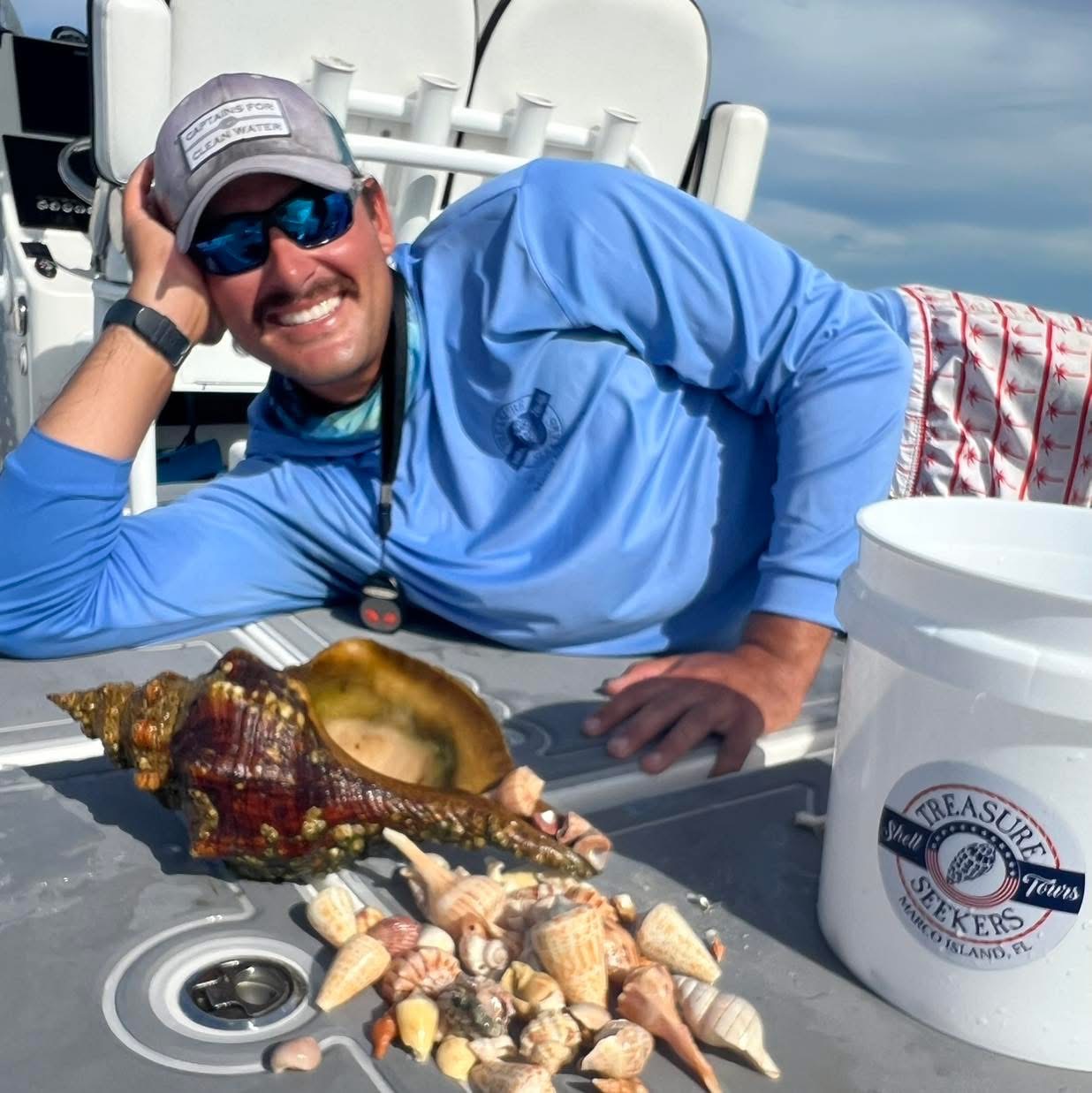 Man in blue shirt and cap smiling with seashells on a boat.