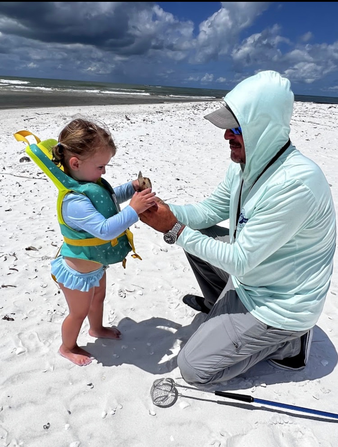 Man and child in swimsuits on beach; man hands child a small object.