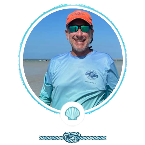 Smiling man in sunglasses and cap at the beach, wearing a light blue shirt.