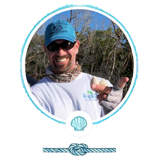 Man in sunglasses and blue cap smiling, holding seashells outdoors.