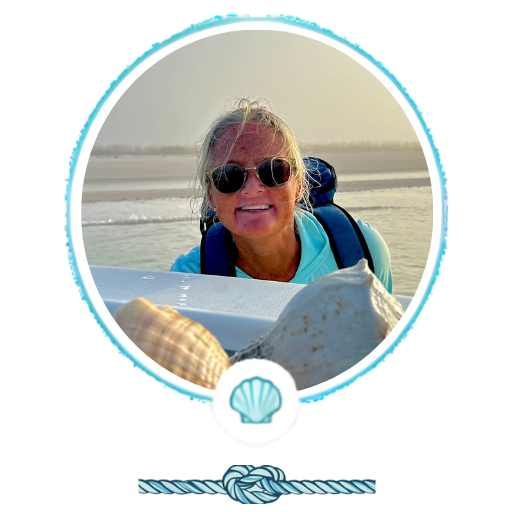 Person smiling on a beach with seashells in foreground, wearing sunglasses and a blue shirt.