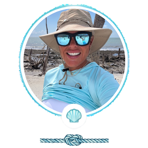Person in hat and sunglasses smiling at beach, with ocean and driftwood in background.