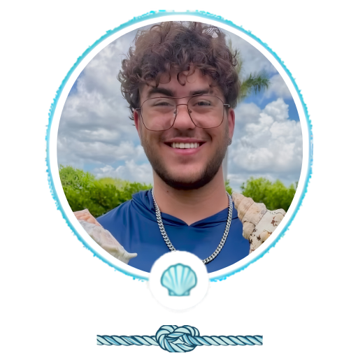 Smiling person with glasses outdoors, wearing blue shirt and chain necklace.