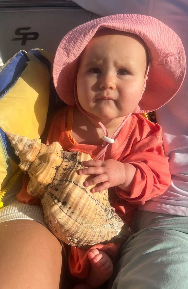 A baby holding a big shell wearing a pink hat.