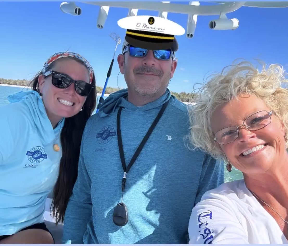Three people smiling on a boat, one wearing a captain's hat.