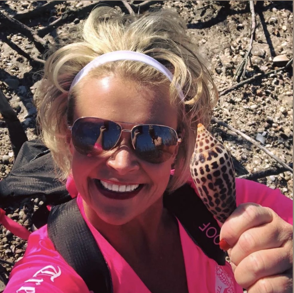 Smiling person in pink shirt holding a spotted shell, wearing sunglasses and a backpack.