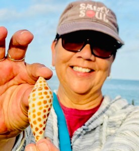 Person holding a spotted seashell on a beach, wearing sunglasses and a cap.