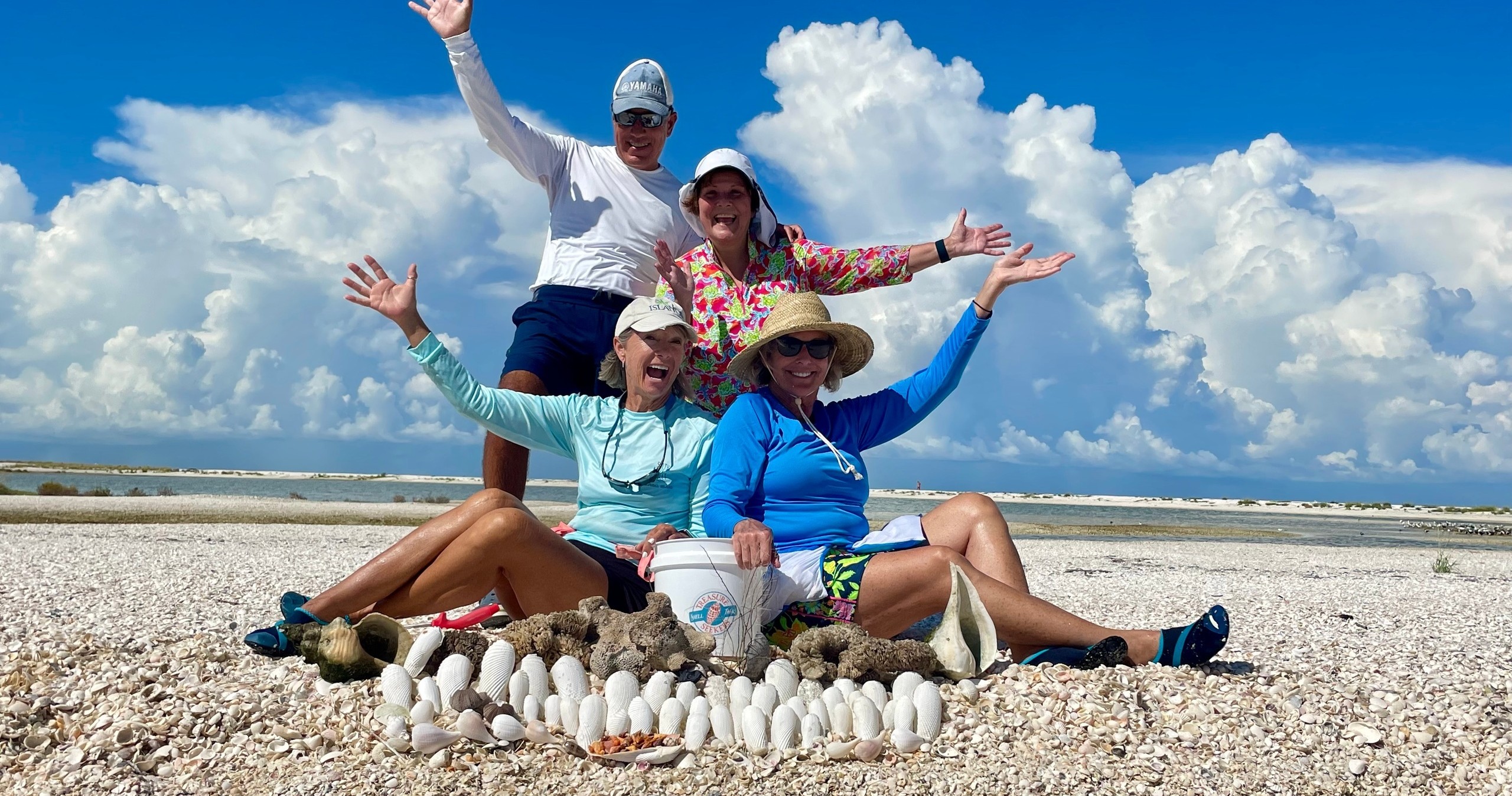 Four people smiling with arms raised, sitting on a shell-covered beach, with cloudy sky in the background.