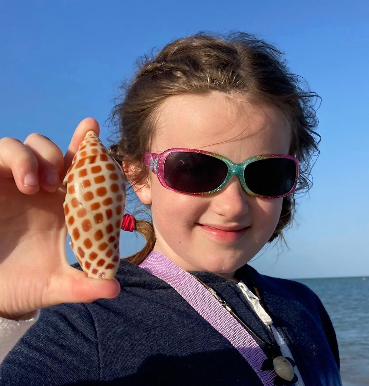 Child with sunglasses holding a patterned seashell by the ocean.