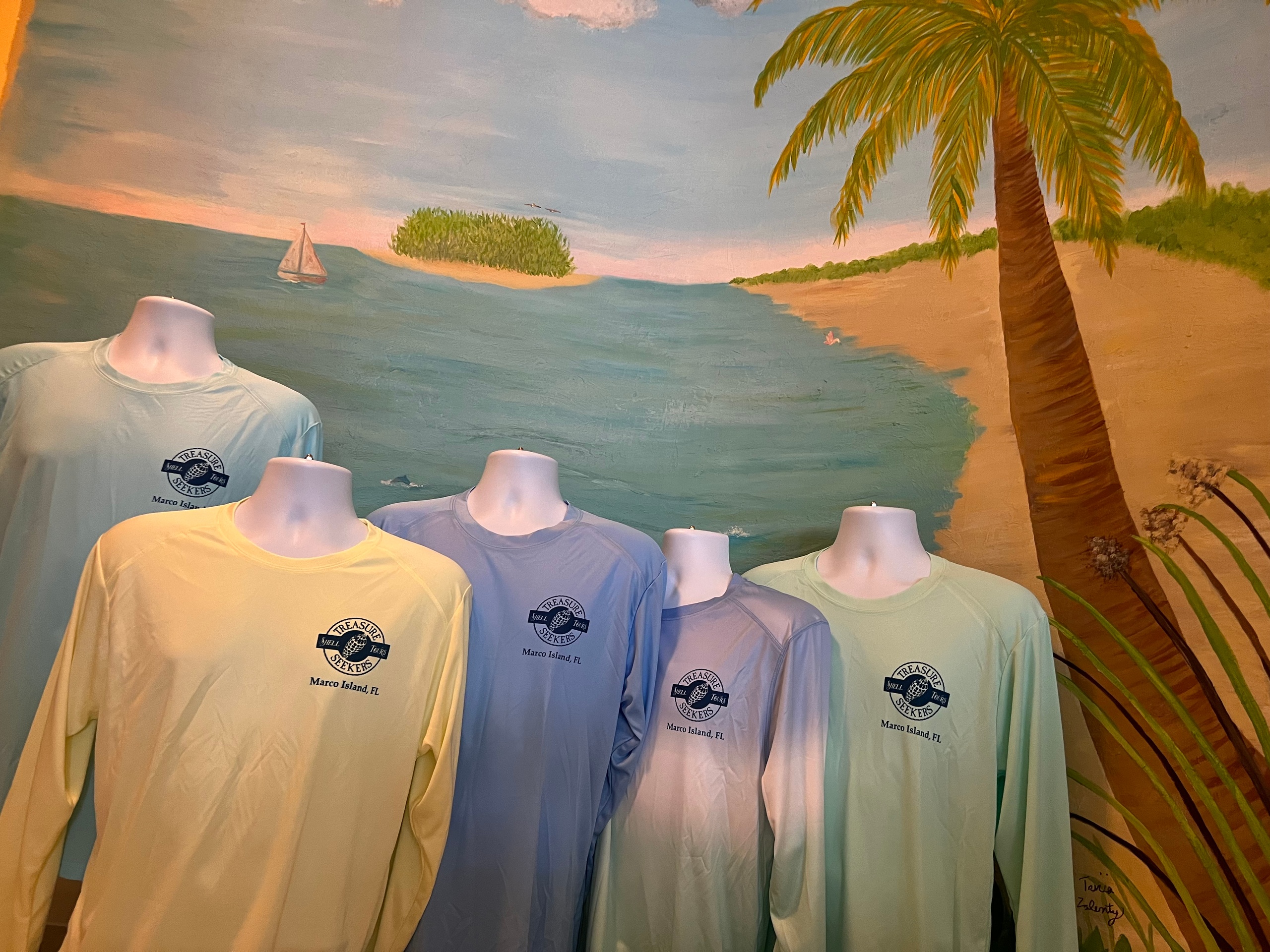 Display of mannequins in long-sleeve shirts with a tropical beach mural background.