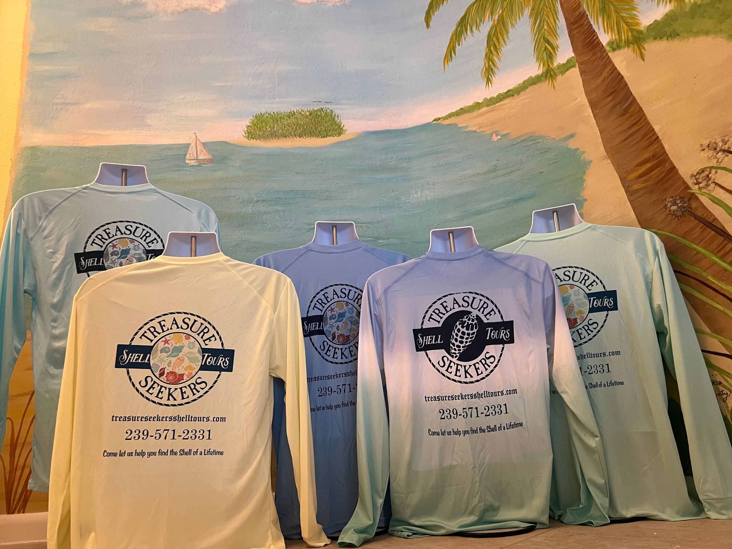 Five mannequins wearing long-sleeve shirts with 'Treasure Seekers' logos in front of a beach mural.