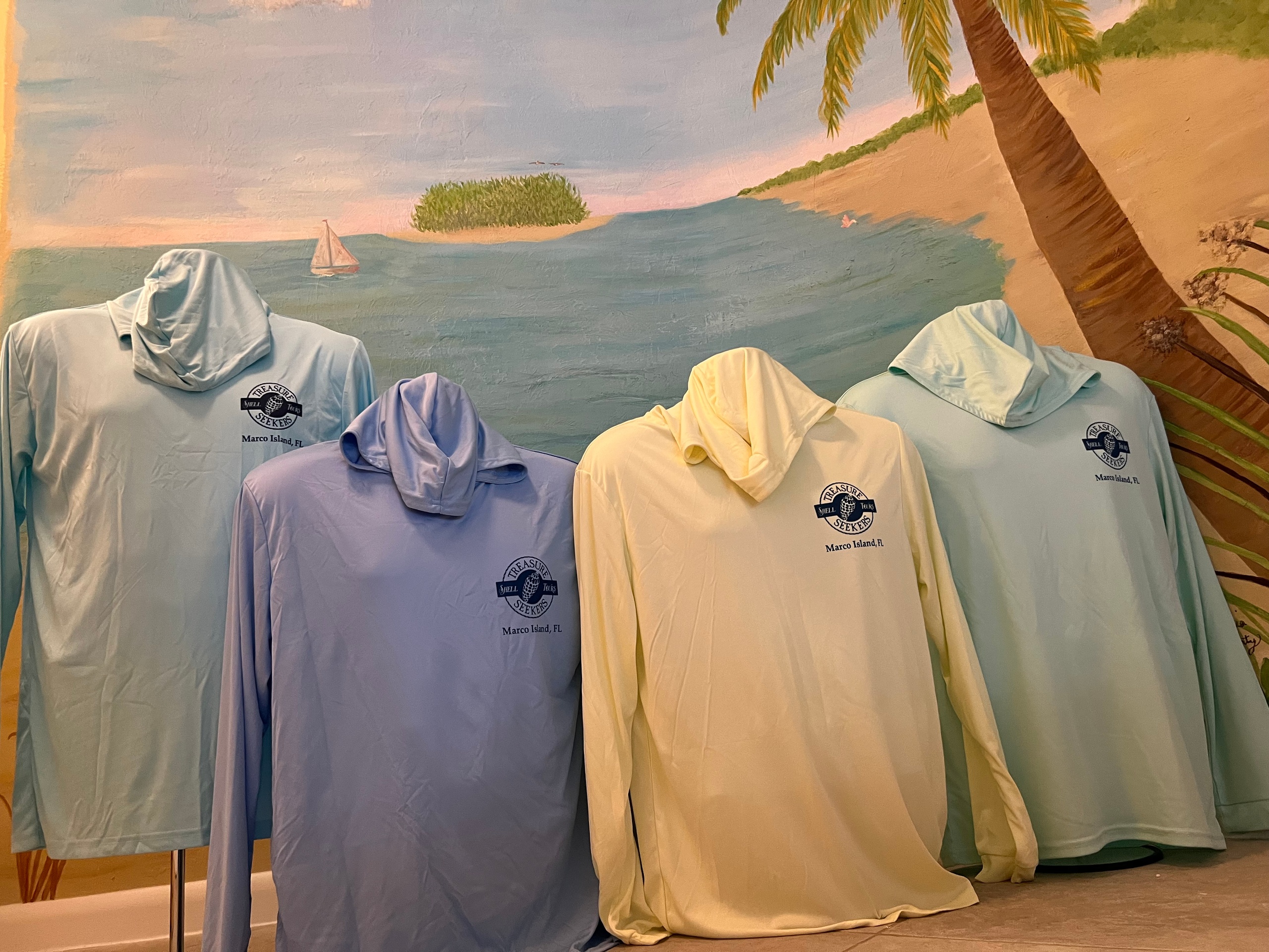 Four hooded shirts on display with a beach mural background.