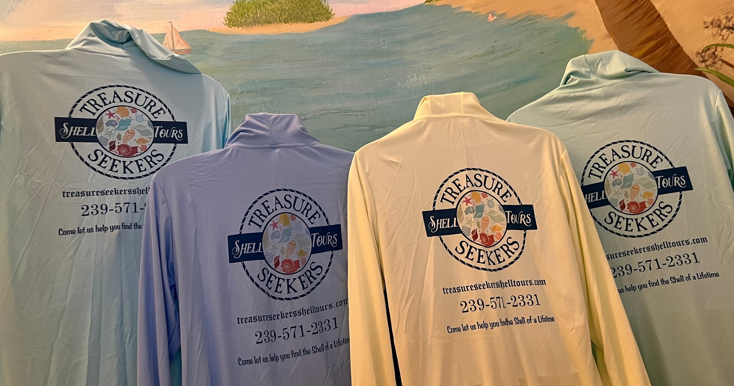 Four Treasure Seekers shirts with logos hanging, beach scene above.