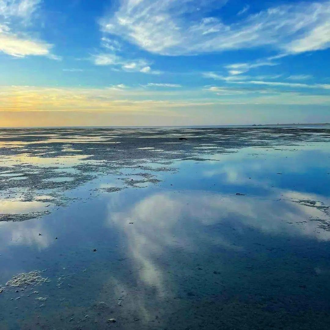 Vast tidal flat with water reflecting blue sky and clouds, horizon visible.