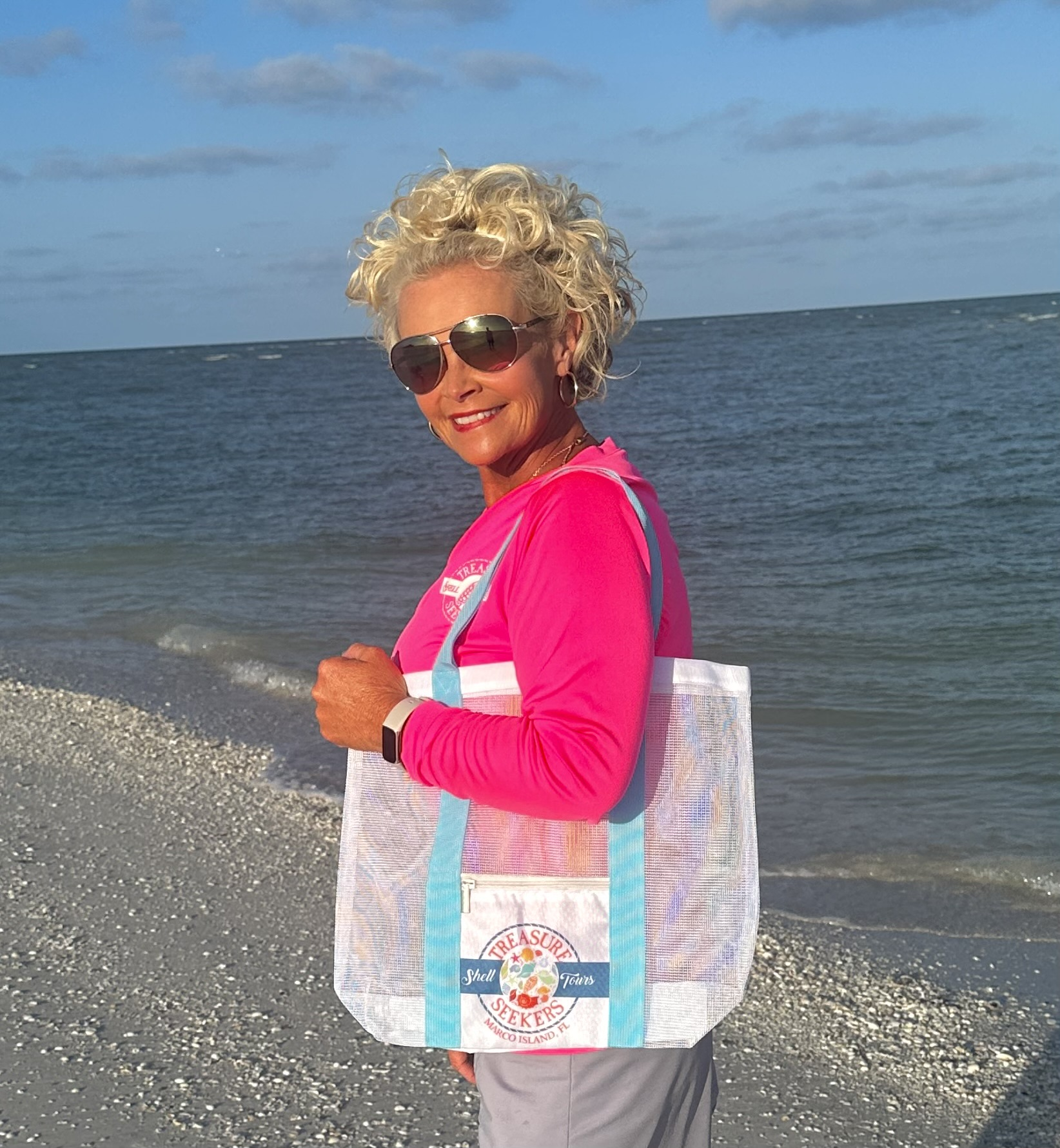 Person with curly hair, sunglasses, pink shirt, and tote bag on beach.